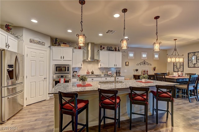 kitchen featuring appliances with stainless steel finishes, wall chimney range hood, an island with sink, decorative light fixtures, and light wood-type flooring