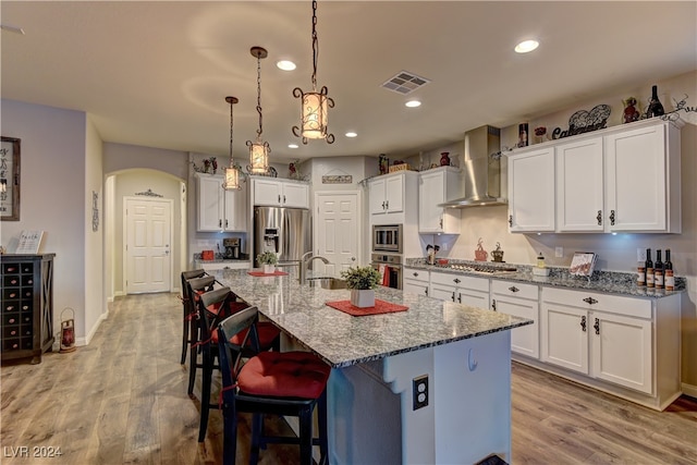 kitchen with wall chimney range hood, a center island with sink, decorative light fixtures, light wood-type flooring, and stainless steel appliances