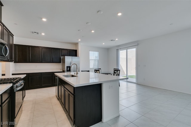 kitchen with dark brown cabinets, a center island with sink, sink, light tile patterned floors, and appliances with stainless steel finishes