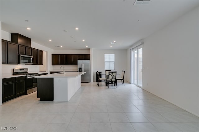 kitchen with an island with sink, appliances with stainless steel finishes, light tile patterned flooring, dark brown cabinetry, and sink