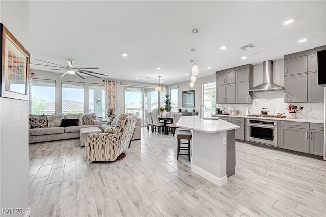 kitchen with wall chimney exhaust hood, sink, stainless steel oven, decorative light fixtures, and an island with sink