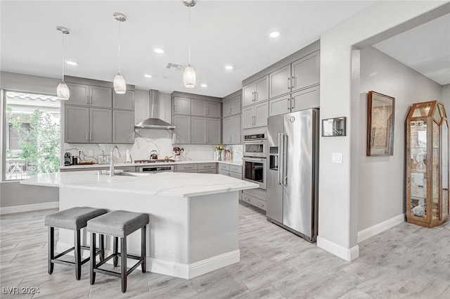 kitchen featuring decorative light fixtures, stainless steel appliances, a kitchen island with sink, decorative backsplash, and wall chimney range hood