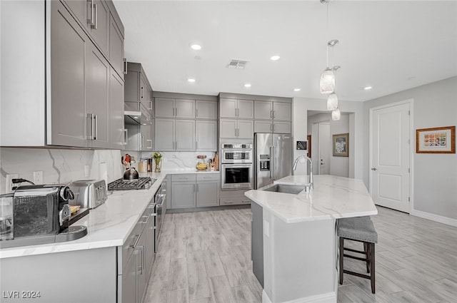 kitchen featuring appliances with stainless steel finishes, pendant lighting, tasteful backsplash, an island with sink, and light stone countertops