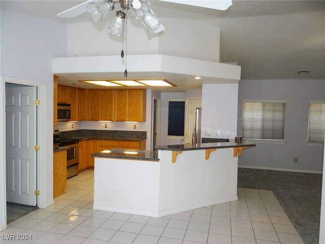 kitchen featuring light carpet, dark stone counters, ceiling fan, appliances with stainless steel finishes, and a breakfast bar area