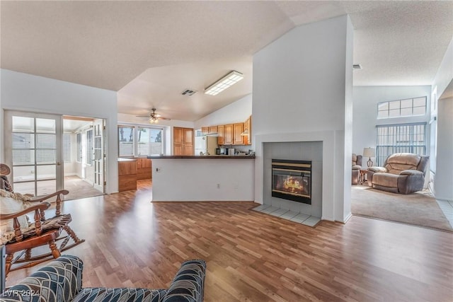 living room with ceiling fan, high vaulted ceiling, a textured ceiling, a tiled fireplace, and light wood-type flooring