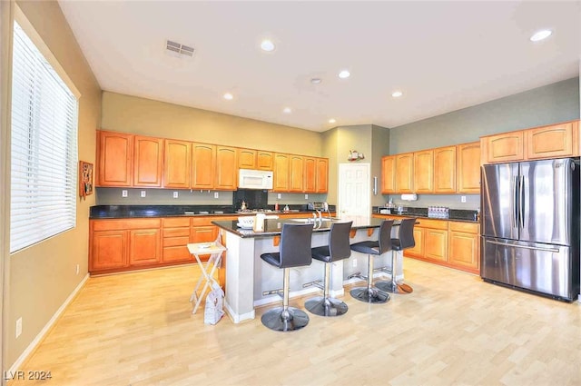 kitchen with dark countertops, white microwave, freestanding refrigerator, an island with sink, and a kitchen breakfast bar