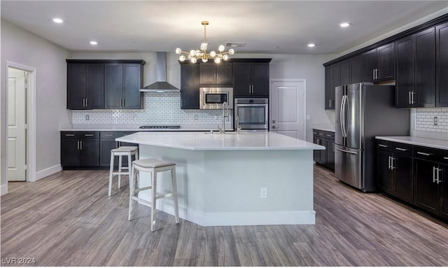 kitchen with wall chimney range hood, a kitchen island with sink, appliances with stainless steel finishes, and light wood-style floors