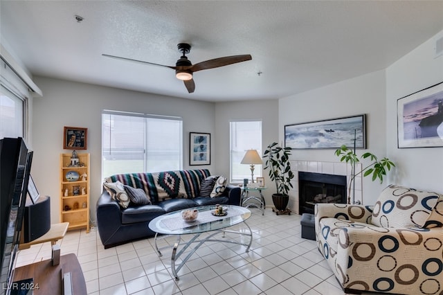 living area featuring ceiling fan, a textured ceiling, a tiled fireplace, and light tile patterned floors