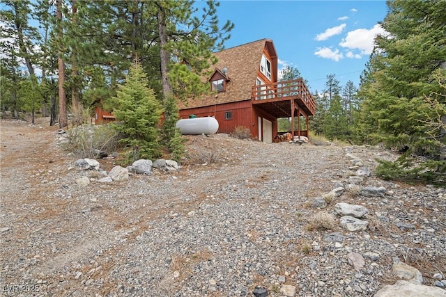 view of property exterior featuring dirt driveway, a deck, a garage, a gambrel roof, and a shingled roof