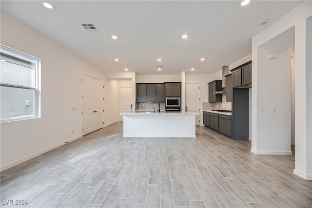 kitchen with gray cabinetry, light wood-type flooring, backsplash, oven, and a kitchen island with sink