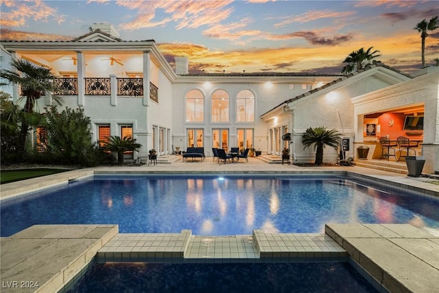 back of house at dusk with a patio, a balcony, ceiling fan, a pool with connected hot tub, and stucco siding