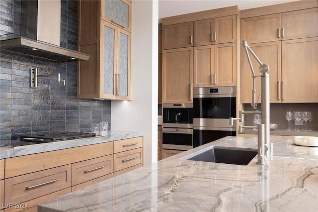 kitchen with light stone counters, gas stovetop, decorative backsplash, double oven, and wall chimney range hood