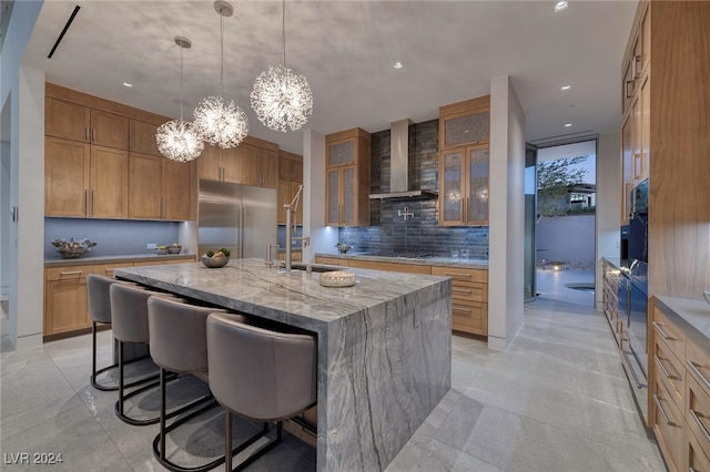 kitchen featuring stainless steel built in fridge, tasteful backsplash, an island with sink, light stone countertops, and wall chimney exhaust hood