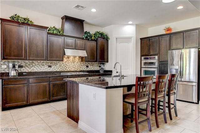 kitchen featuring dark stone countertops, stainless steel appliances, a breakfast bar area, a center island with sink, and tasteful backsplash