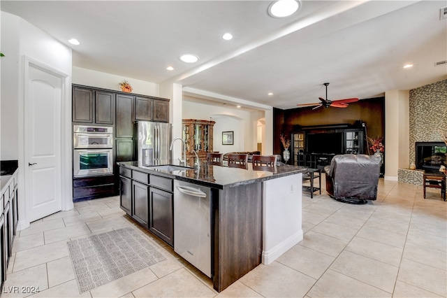 kitchen with dark stone countertops, appliances with stainless steel finishes, sink, ceiling fan, and a kitchen island with sink