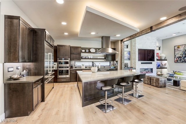 kitchen featuring light hardwood / wood-style flooring, wall chimney range hood, a stone fireplace, a kitchen island, and a breakfast bar