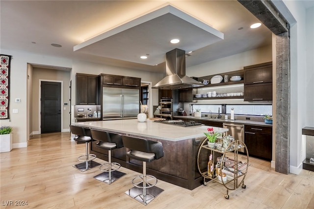 kitchen with appliances with stainless steel finishes, dark brown cabinets, island exhaust hood, a kitchen breakfast bar, and a kitchen island