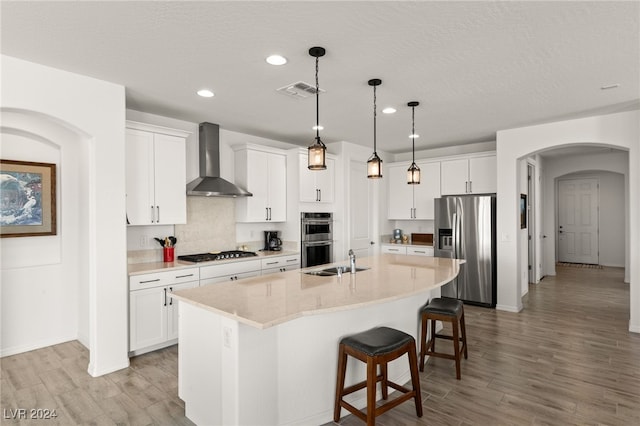 kitchen featuring pendant lighting, wall chimney range hood, appliances with stainless steel finishes, a kitchen island with sink, and white cabinets