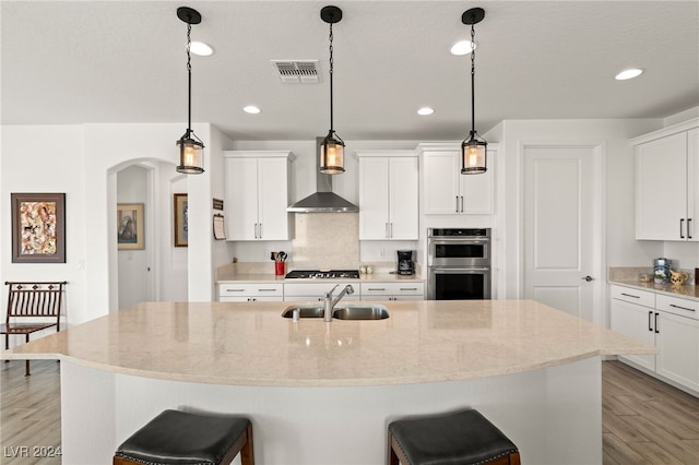 kitchen with sink, white cabinetry, stainless steel double oven, a center island with sink, and wall chimney exhaust hood