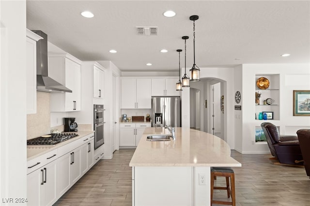 kitchen with appliances with stainless steel finishes, hanging light fixtures, white cabinets, a center island with sink, and wall chimney exhaust hood