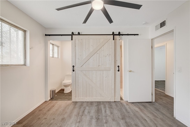 unfurnished bedroom with ceiling fan, a barn door, light wood-type flooring, and connected bathroom