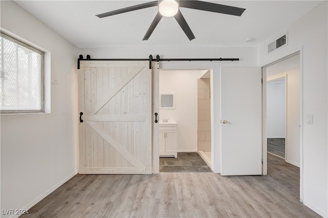 unfurnished bedroom featuring wood-type flooring, a barn door, ensuite bathroom, and ceiling fan