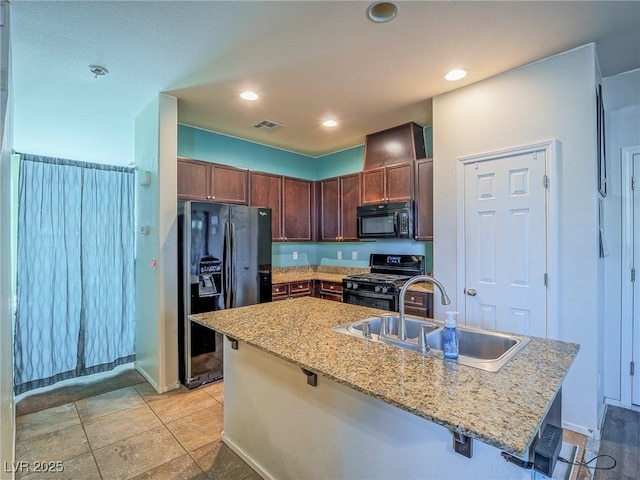 kitchen featuring a kitchen island with sink, black appliances, sink, light stone counters, and a kitchen bar