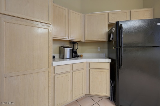 kitchen featuring light brown cabinets, light tile patterned floors, tile counters, and black fridge