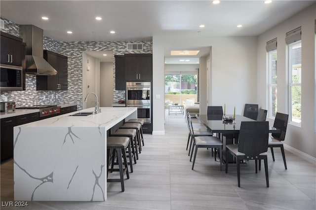 kitchen with appliances with stainless steel finishes, sink, wall chimney exhaust hood, and a center island with sink