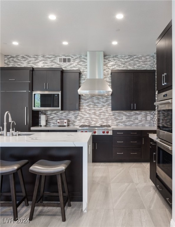 kitchen featuring built in appliances, a breakfast bar area, sink, a kitchen island with sink, and wall chimney range hood