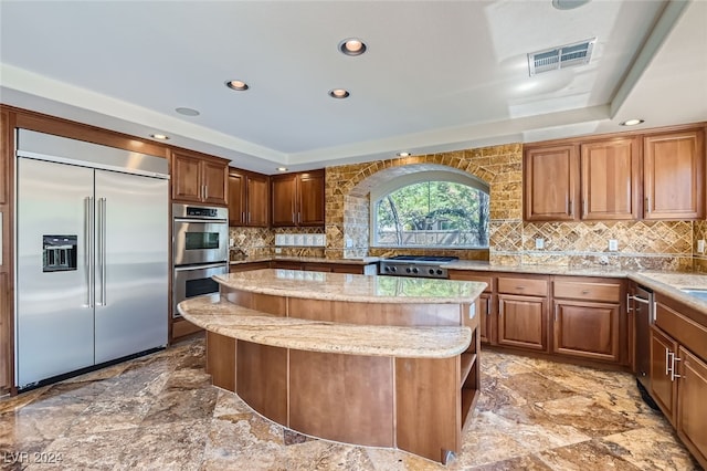 kitchen with stainless steel appliances, light stone counters, a raised ceiling, backsplash, and a kitchen island