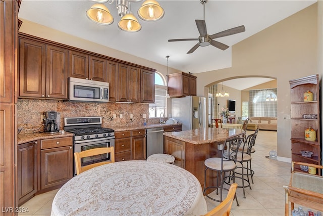 kitchen with ceiling fan with notable chandelier, stainless steel appliances, light stone countertops, and a center island