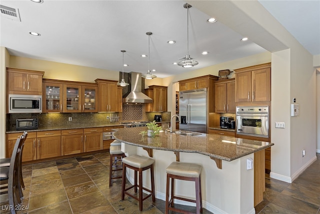 kitchen featuring a center island with sink, built in appliances, hanging light fixtures, wall chimney range hood, and backsplash