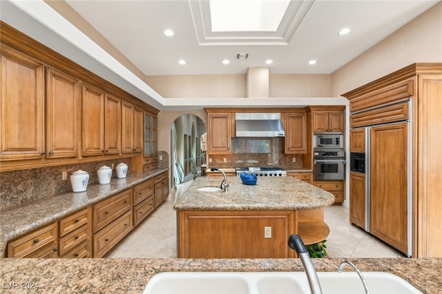 kitchen featuring built in appliances, an island with sink, sink, light stone counters, and wall chimney range hood