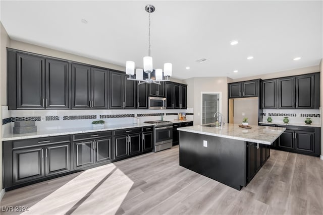 kitchen with a kitchen island with sink, pendant lighting, light wood-type flooring, backsplash, and stainless steel appliances