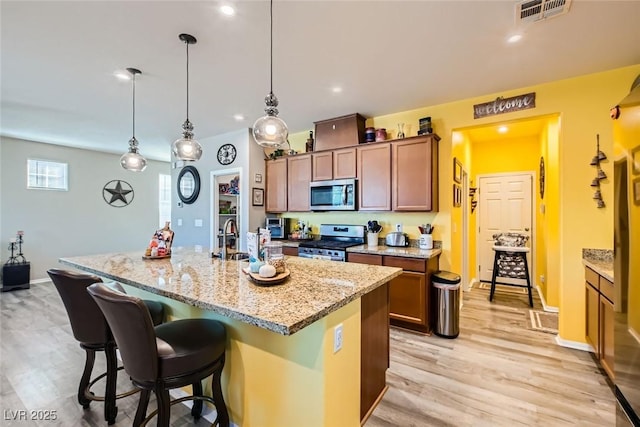 kitchen featuring hanging light fixtures, a kitchen island with sink, light stone counters, stainless steel appliances, and light wood-type flooring