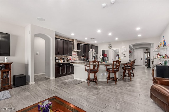 kitchen with a kitchen breakfast bar, light stone counters, wall chimney exhaust hood, a center island with sink, and dark brown cabinetry