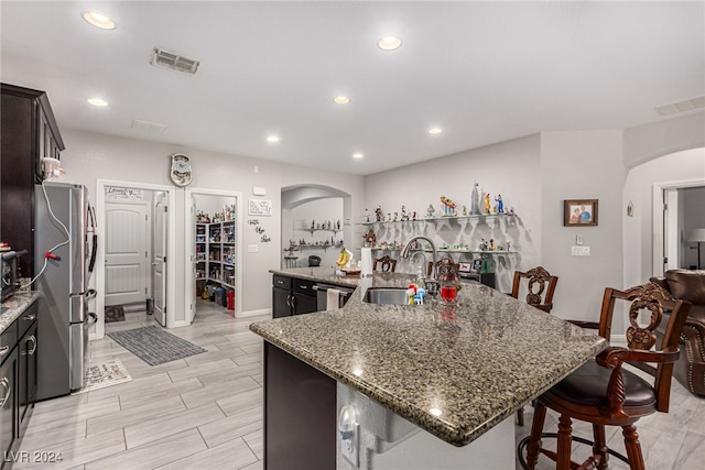 kitchen with an island with sink, a breakfast bar, dark stone countertops, and stainless steel fridge