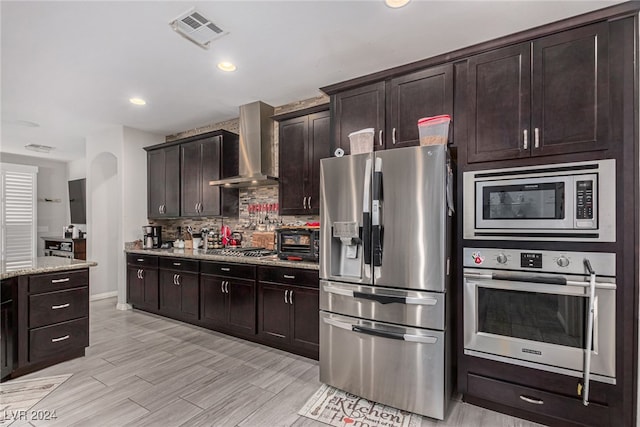 kitchen featuring light wood-type flooring, appliances with stainless steel finishes, light stone counters, wall chimney range hood, and dark brown cabinets