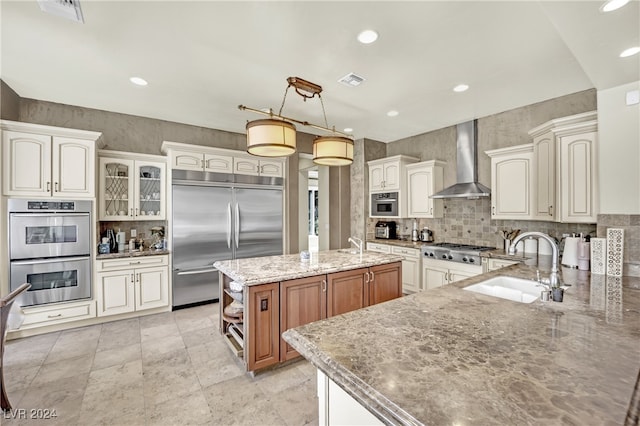 kitchen featuring light stone counters, an island with sink, sink, wall chimney exhaust hood, and appliances with stainless steel finishes