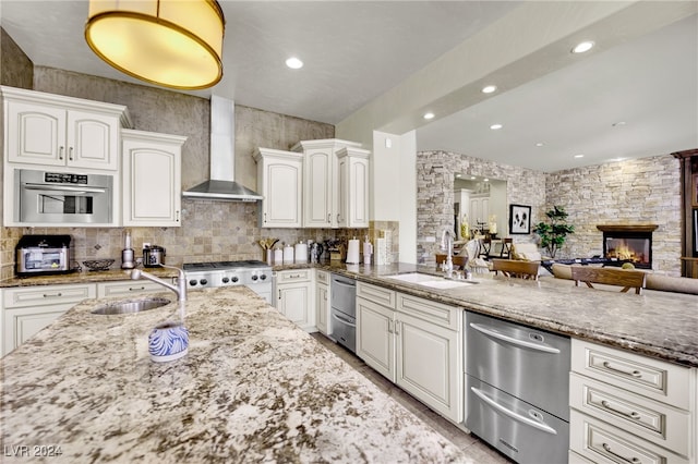 kitchen featuring sink, white cabinets, wall chimney exhaust hood, appliances with stainless steel finishes, and a stone fireplace