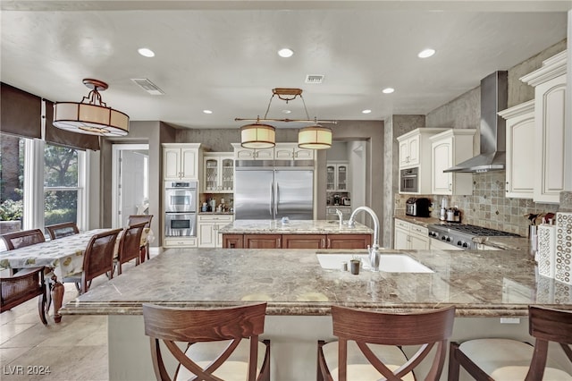 kitchen featuring pendant lighting, stainless steel appliances, an island with sink, and wall chimney range hood