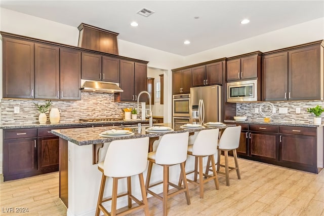 kitchen featuring dark stone countertops, stainless steel appliances, light wood-type flooring, and a kitchen island with sink