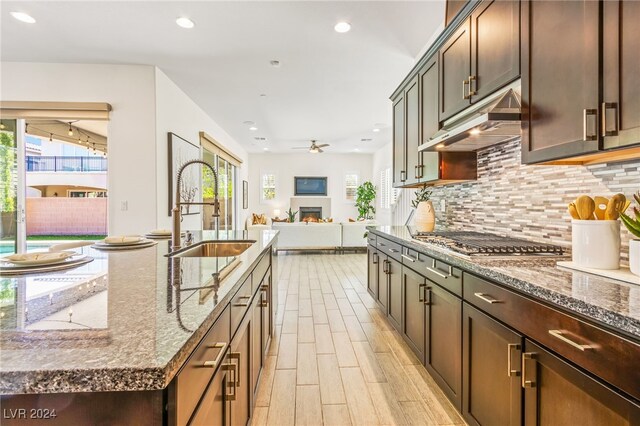 kitchen with an island with sink, stone counters, stainless steel gas cooktop, sink, and light hardwood / wood-style floors