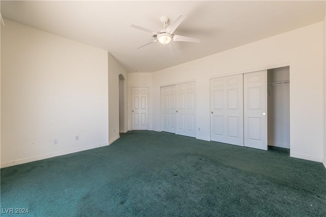 unfurnished bedroom with two closets, a ceiling fan, dark carpet, arched walkways, and baseboards