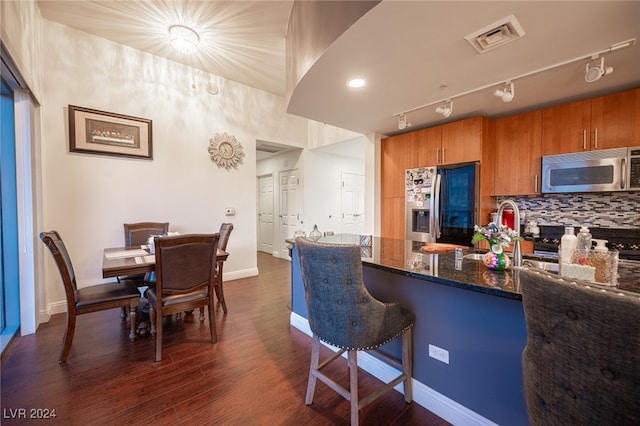 kitchen featuring track lighting, dark wood-type flooring, stainless steel appliances, a breakfast bar, and dark stone counters