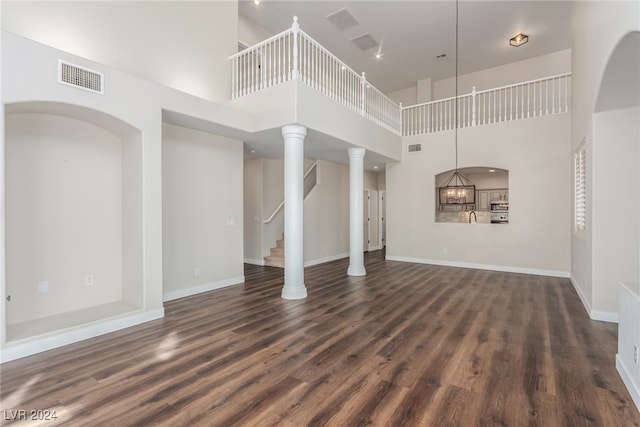 unfurnished living room featuring a towering ceiling, an inviting chandelier, dark hardwood / wood-style floors, and ornate columns