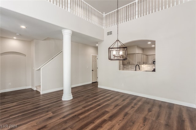 unfurnished living room featuring a towering ceiling, an inviting chandelier, decorative columns, dark hardwood / wood-style floors, and sink