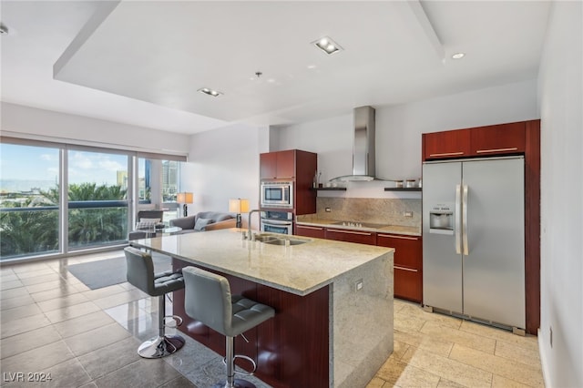 kitchen featuring light stone counters, ventilation hood, a kitchen island with sink, a breakfast bar, and appliances with stainless steel finishes