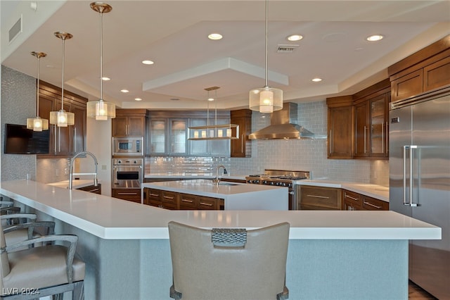 kitchen featuring pendant lighting, wall chimney range hood, built in appliances, a kitchen breakfast bar, and decorative backsplash